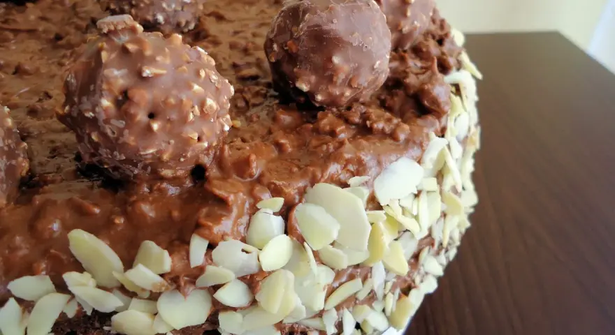 Pyszny tort Ferrero Rocher, oblany czekoladą i obsypany płatkami migdałów, z całymi czekoladkami na wierzchu.