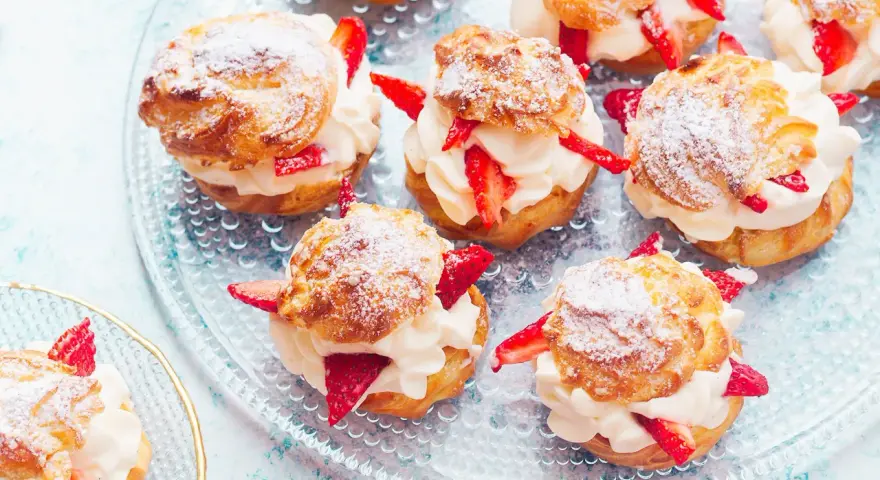 Pyszne ptysie z kremem patissiere i świeżymi truskawkami, posypane cukrem pudrem, prezentują się apetycznie na szklanej paterze.