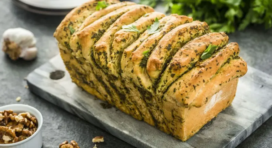Pyszny chlebek rwany z pesto, udekorowany listkami bazylii, prezentuje się apetycznie na kamiennej desce. Obok czosnek i orzechy włoskie.