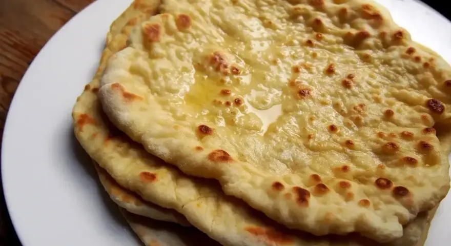 Stos świeżo upieczonych, lekko przypieczonych placków roti i naan, polanych roztopionym masłem, na białym talerzu.