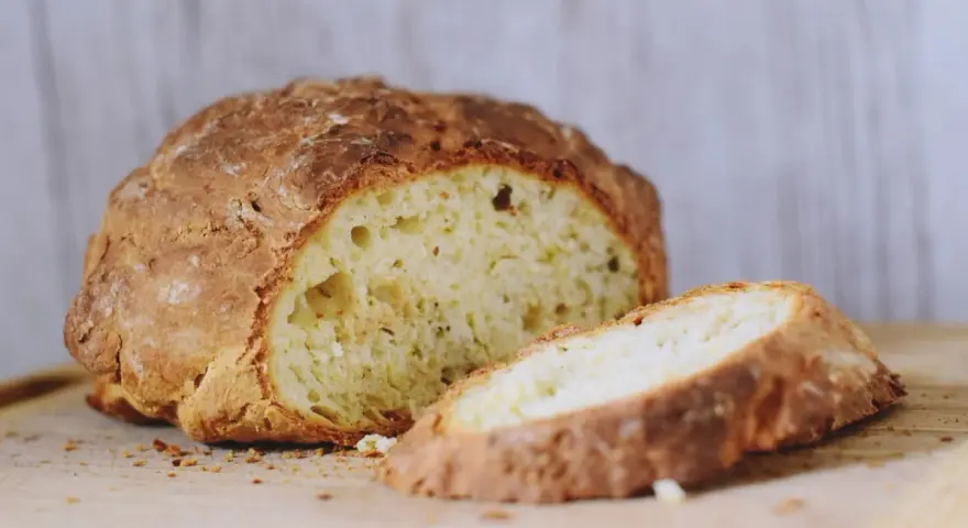 Chrupiący, złocisty bochenek soda bread, z jednym plastrem odciętym, na drewnianej desce.