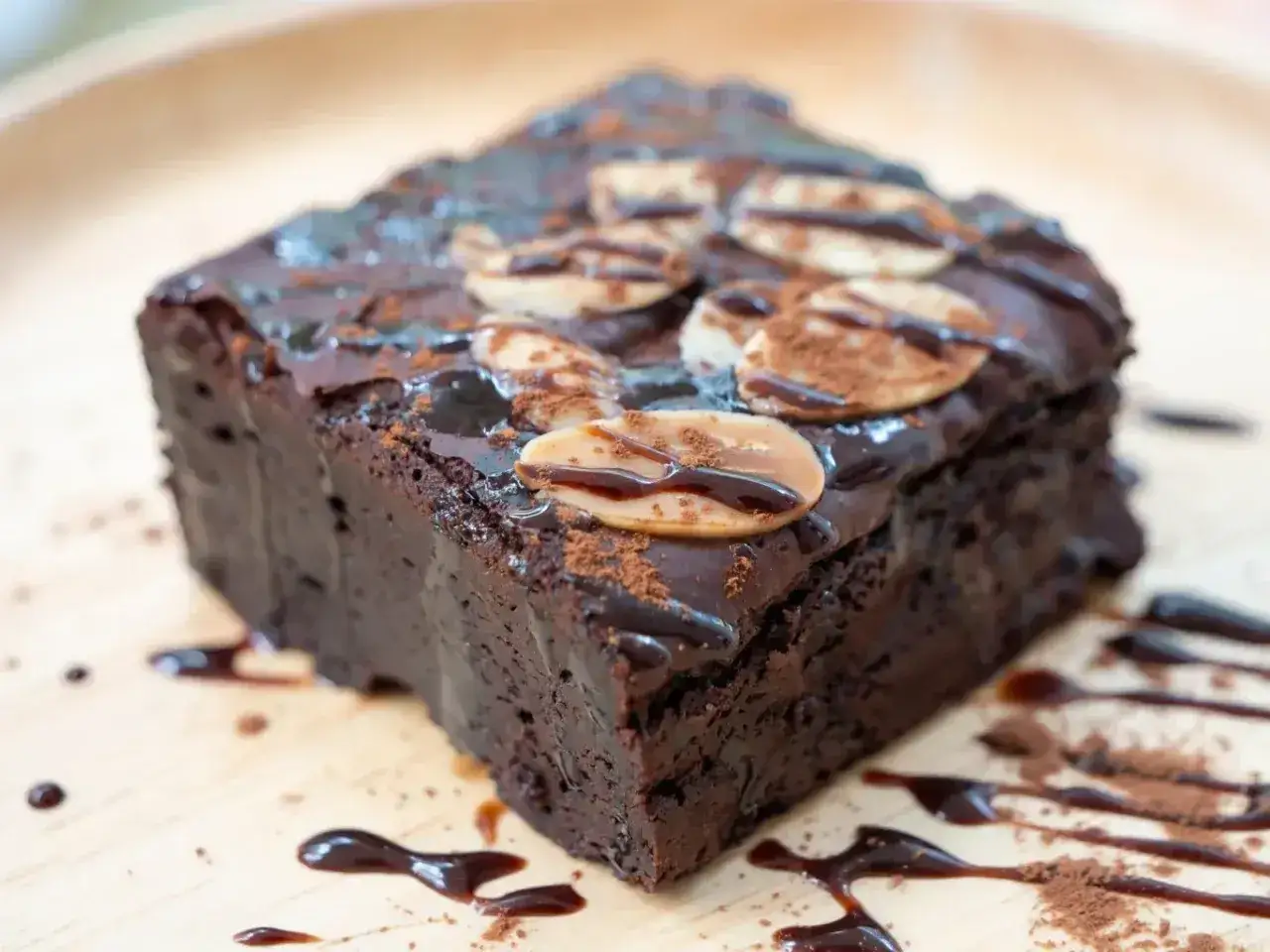 Pyszne brownie bezglutenowe, polane czekoladą i posypane płatkami migdał&oacute;w, na drewnianej desce.