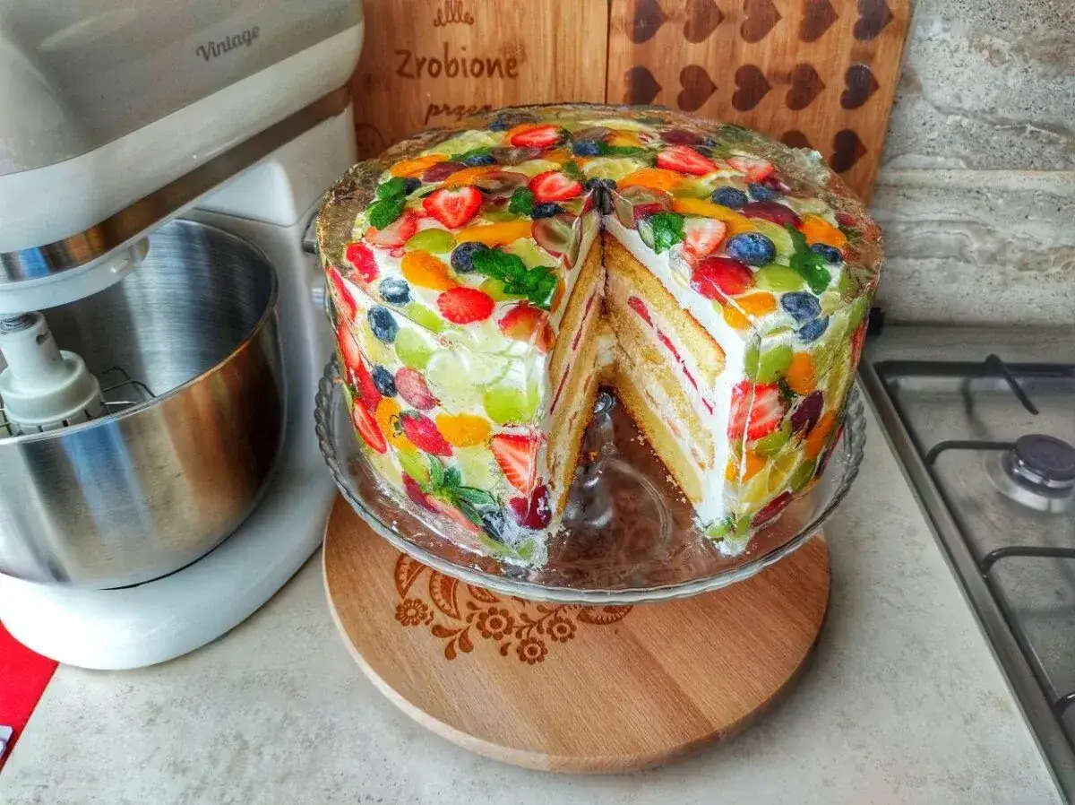 Kolorowy tort z galaretką i owocami, z wyciętym kawałkiem, prezentuje się apetycznie.