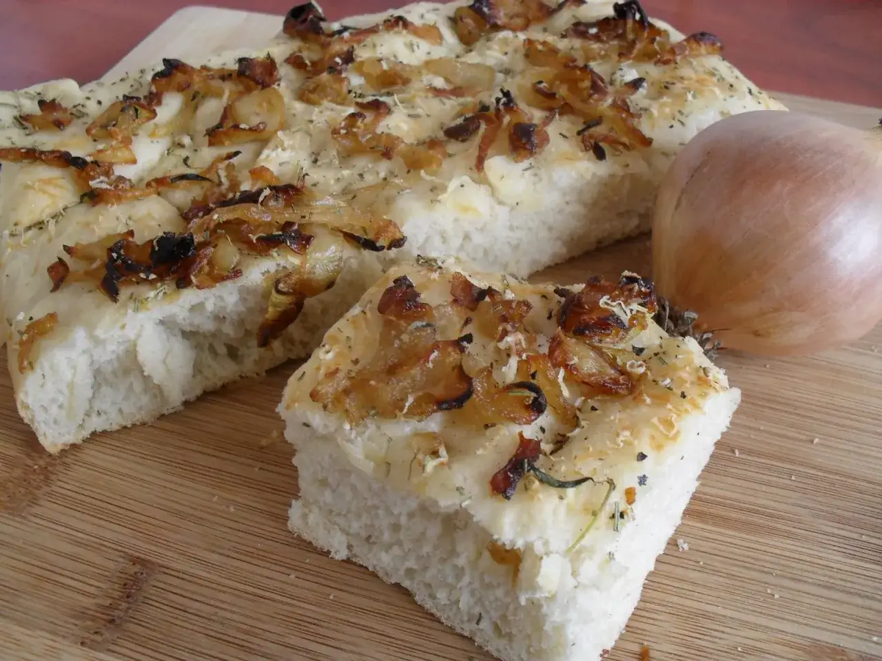 Puszysta focaccia z cebulą, posypana ziołami i startym serem. Obok leży cała cebula, sugerując jej obecność w tym pysznym wypieku.