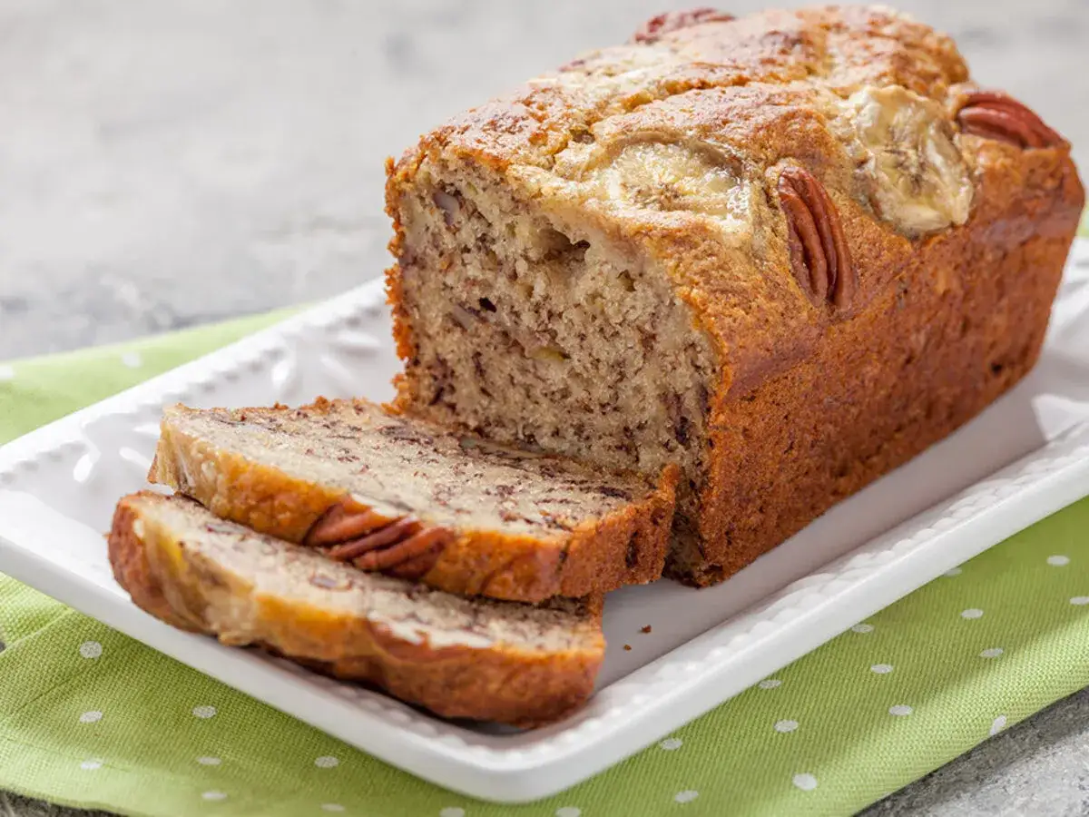 Pyszny chlebek bananowy z orzechami pekan i plasterkami banana na wierzchu, pokrojony na kromki, na białej paterze z zieloną serwetką.