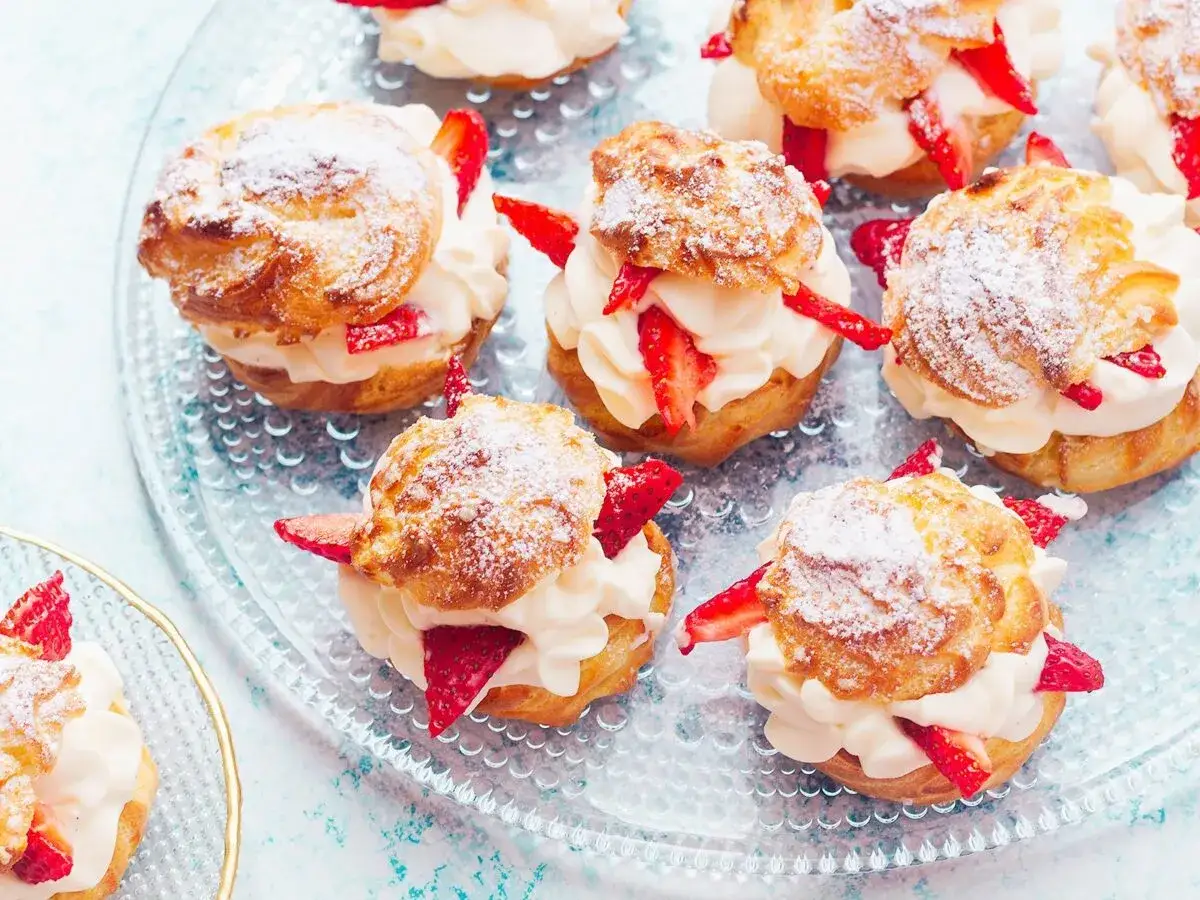 Pyszne ptysie z kremem patissiere i świeżymi truskawkami, posypane cukrem pudrem, kuszą na szklanej tacy.
